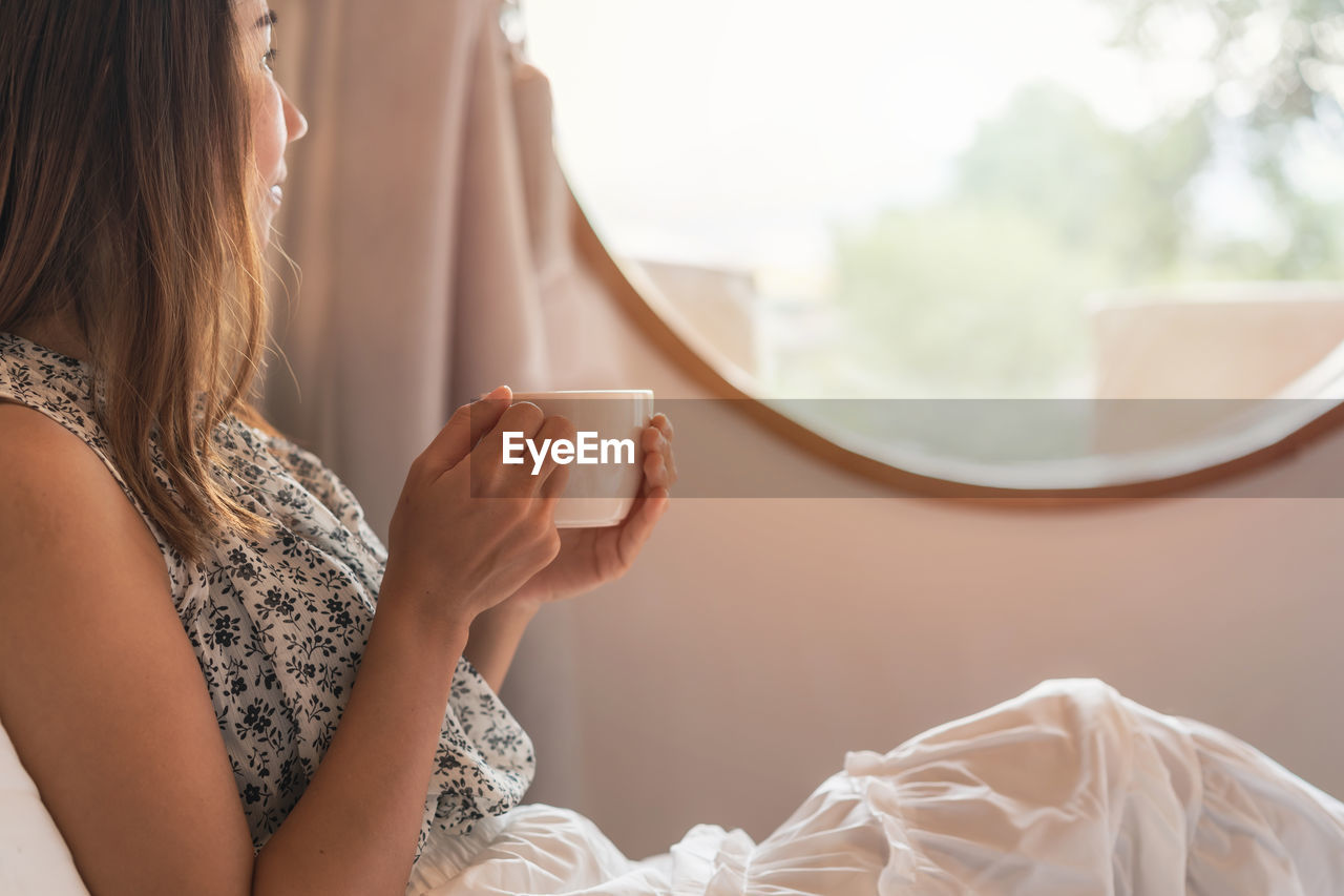 Woman holding coffee cup while relaxing at bed