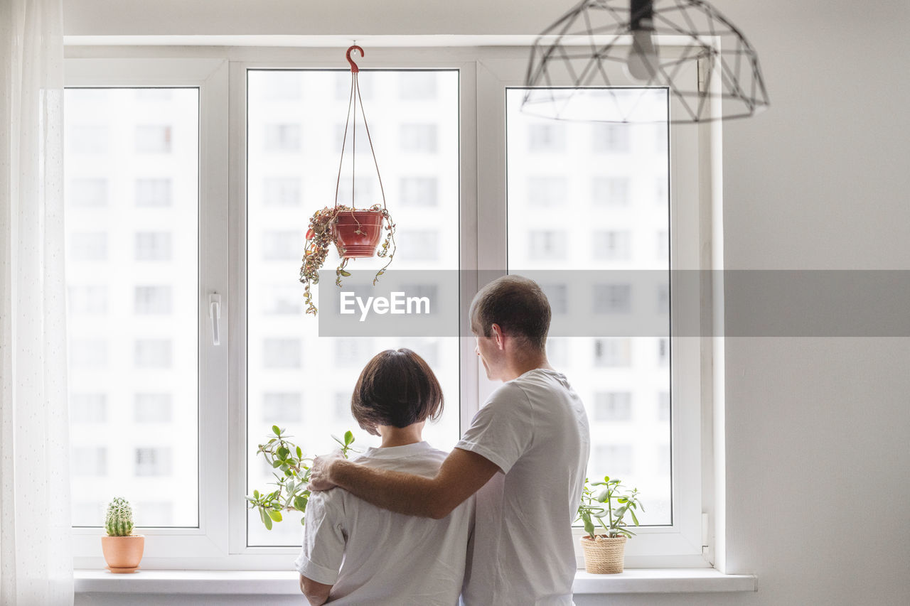 Husband and wife embracing while looking through window at home