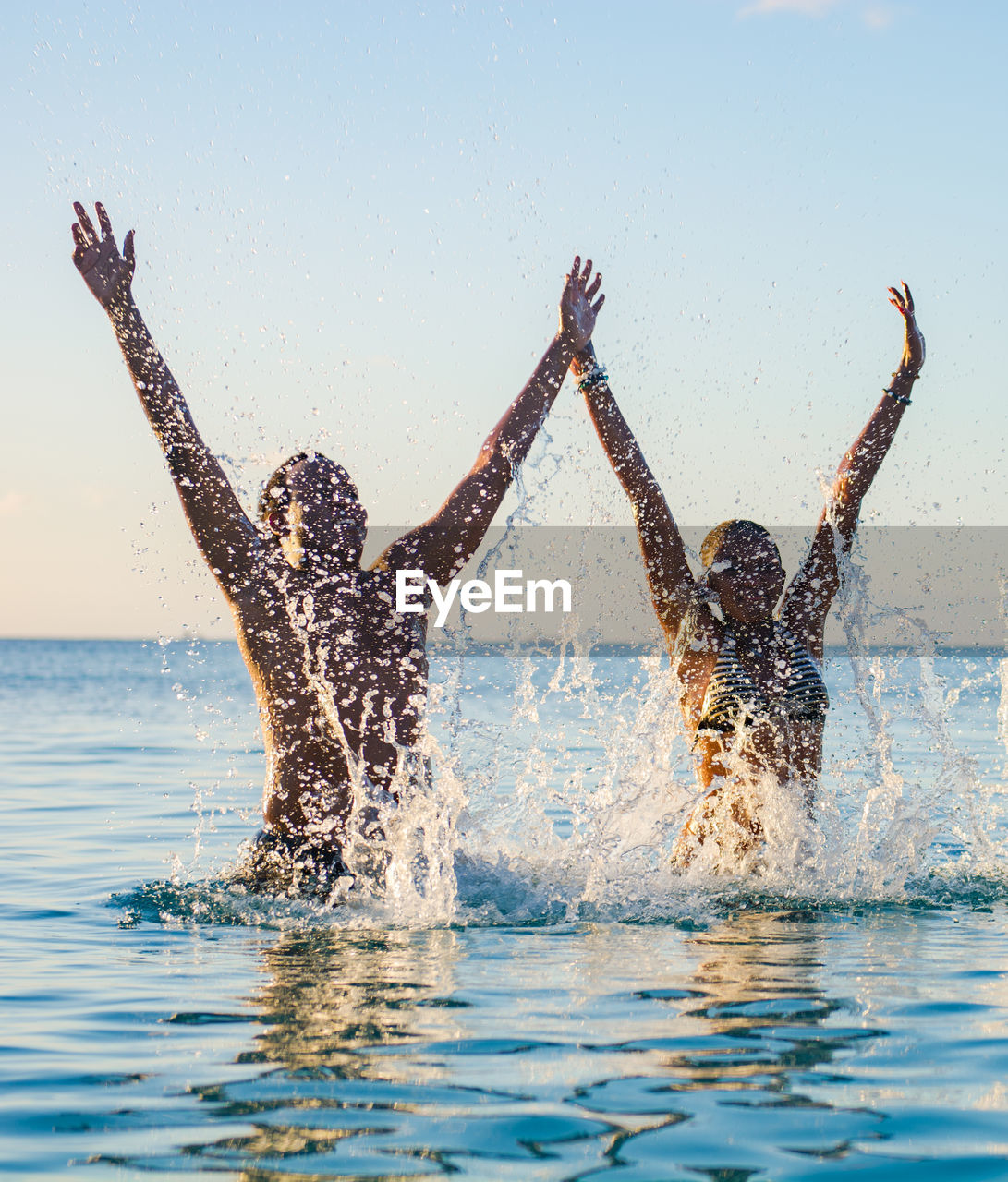 Portrait of two people emerging from underwater with hands raised high 