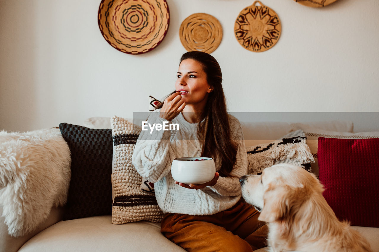 Woman eating food while sitting by dog on sofa