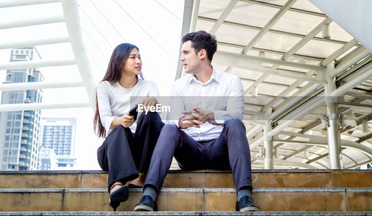 Low angle view of couple talking while sitting on steps in city