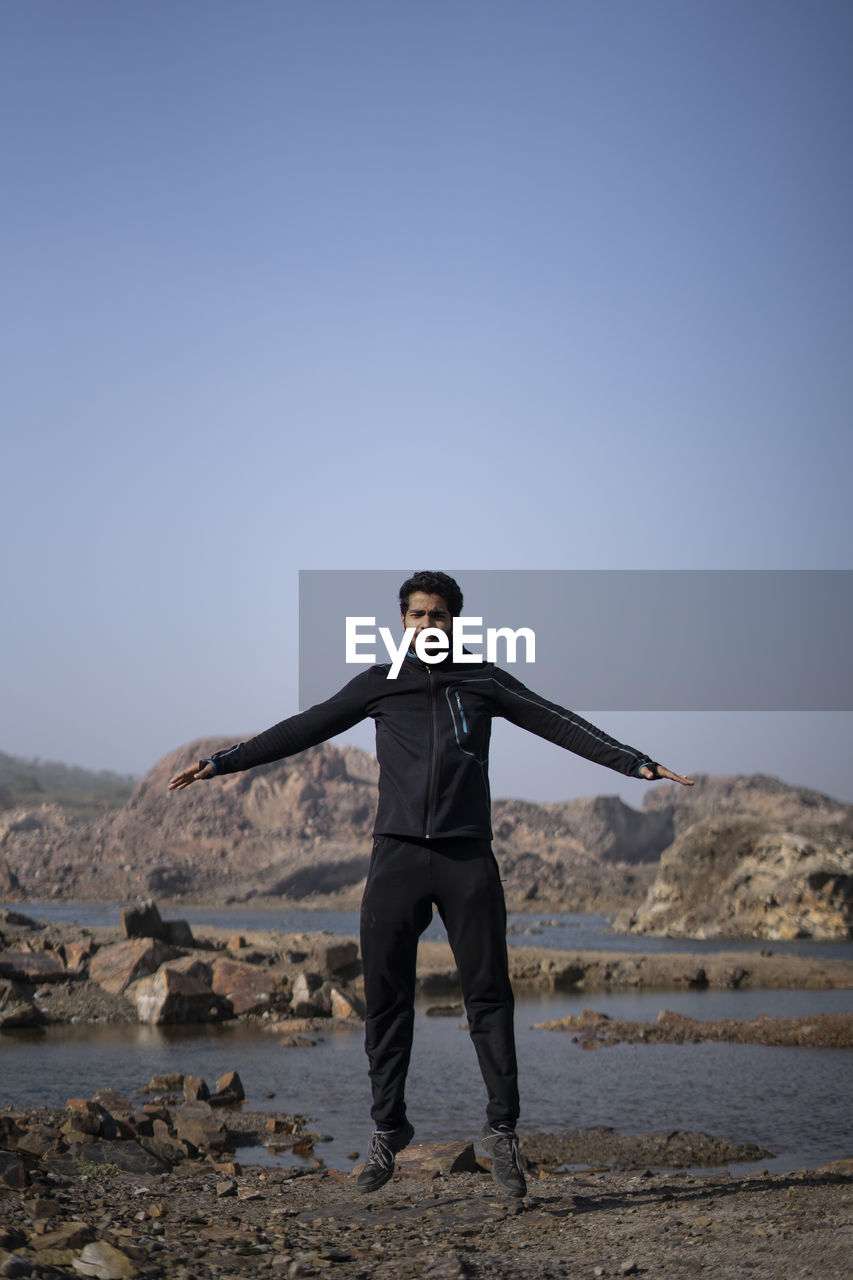 Young athletic man doing burpee near a lake surrounded with mountains early in the morning.