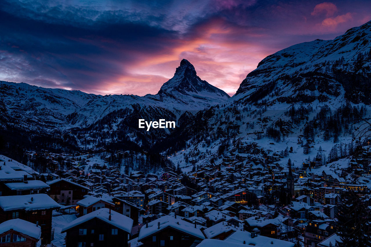 Snow covered houses and mountains against sky during sunset