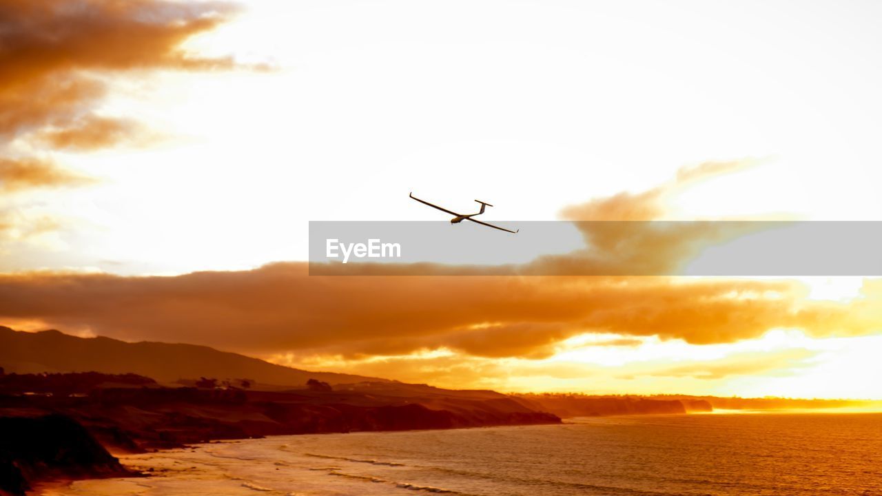 LOW ANGLE VIEW OF AIRPLANE FLYING OVER SEA AGAINST SKY DURING SUNSET