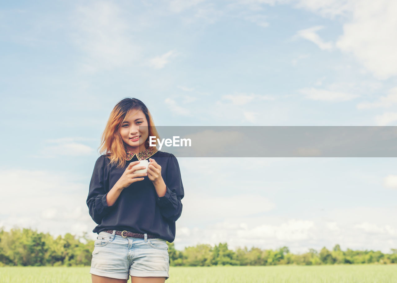 Smiling young woman with coffee cup standing against sky