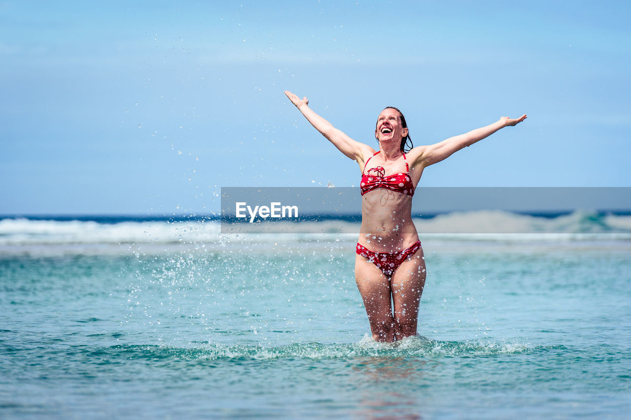 Happy mid adult woman with arms outstretched in sea against sky