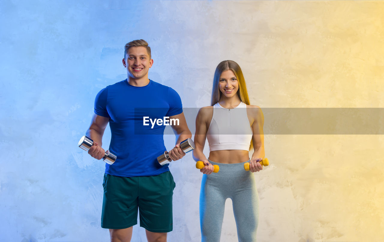 Portrait of young couple holding dumbbell standing against illuminated wall