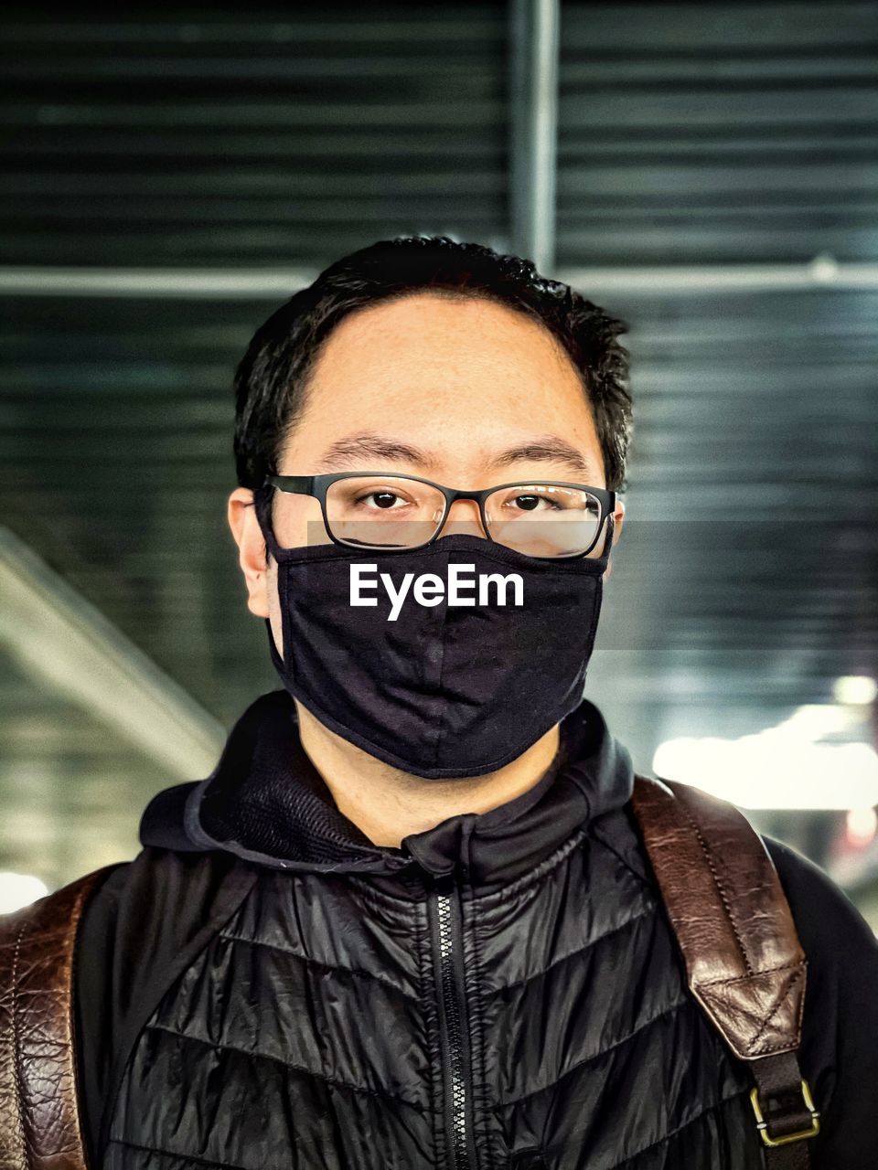 Portrait of young man wearing eyeglasses and black face mask against ceiling and lights.