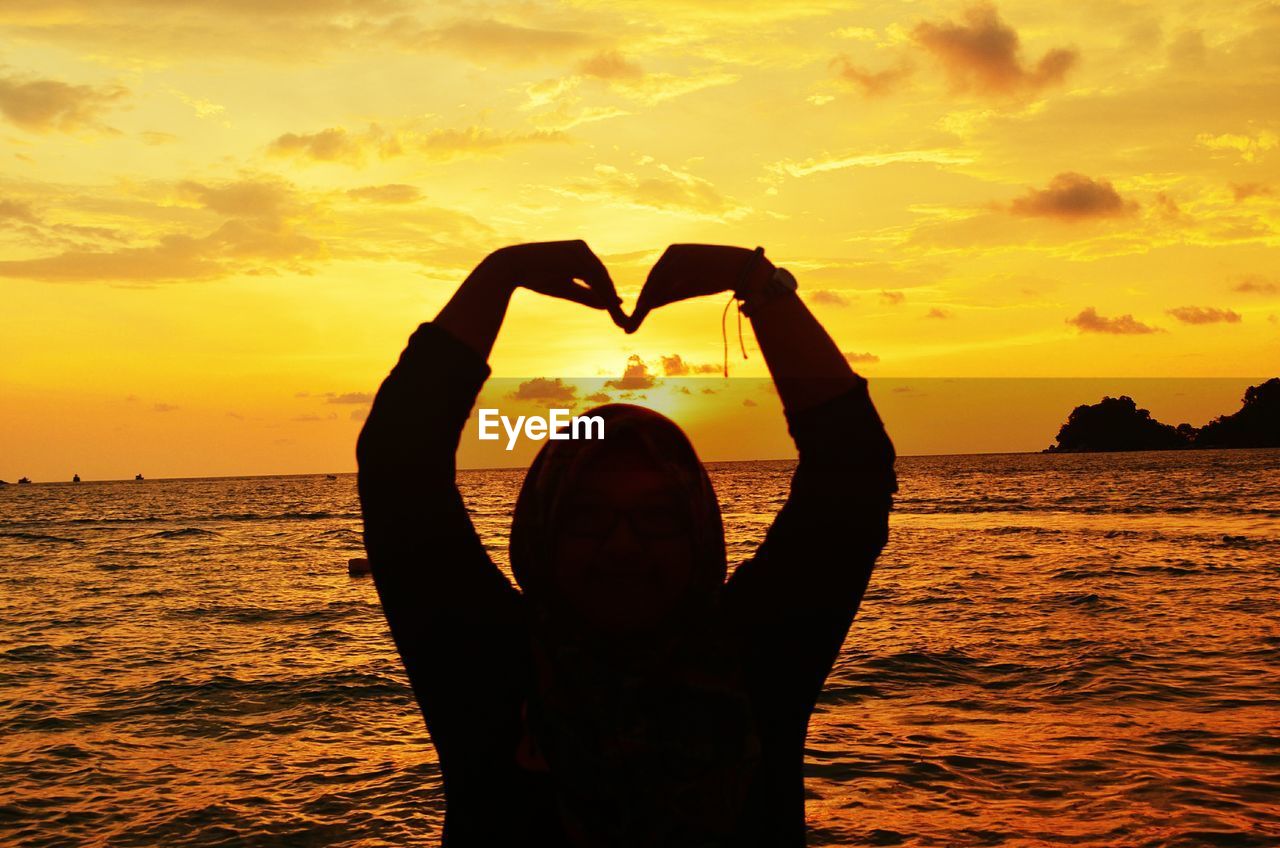 Silhouette man forming heart shape while standing against sea during sunset