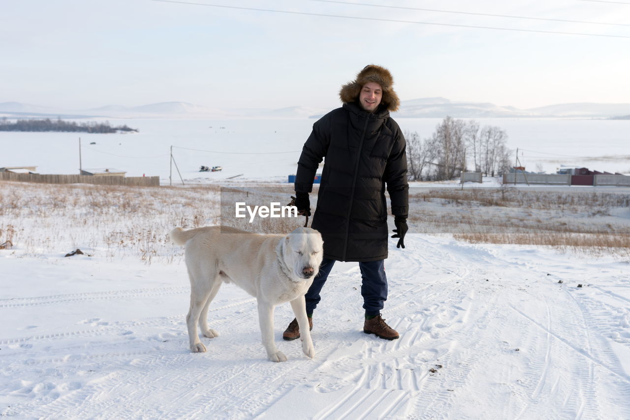 A young man stands with a big dog on a rural road amid a frozen lake in winter.