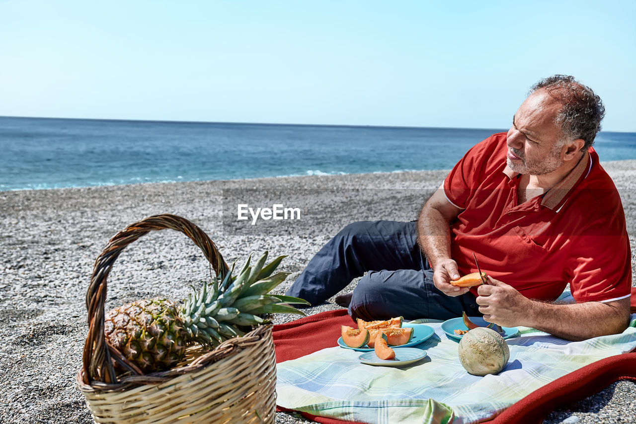 Middle aged couple having a picnic at the seaside with fresh exotic fruit. man cuts a melon.