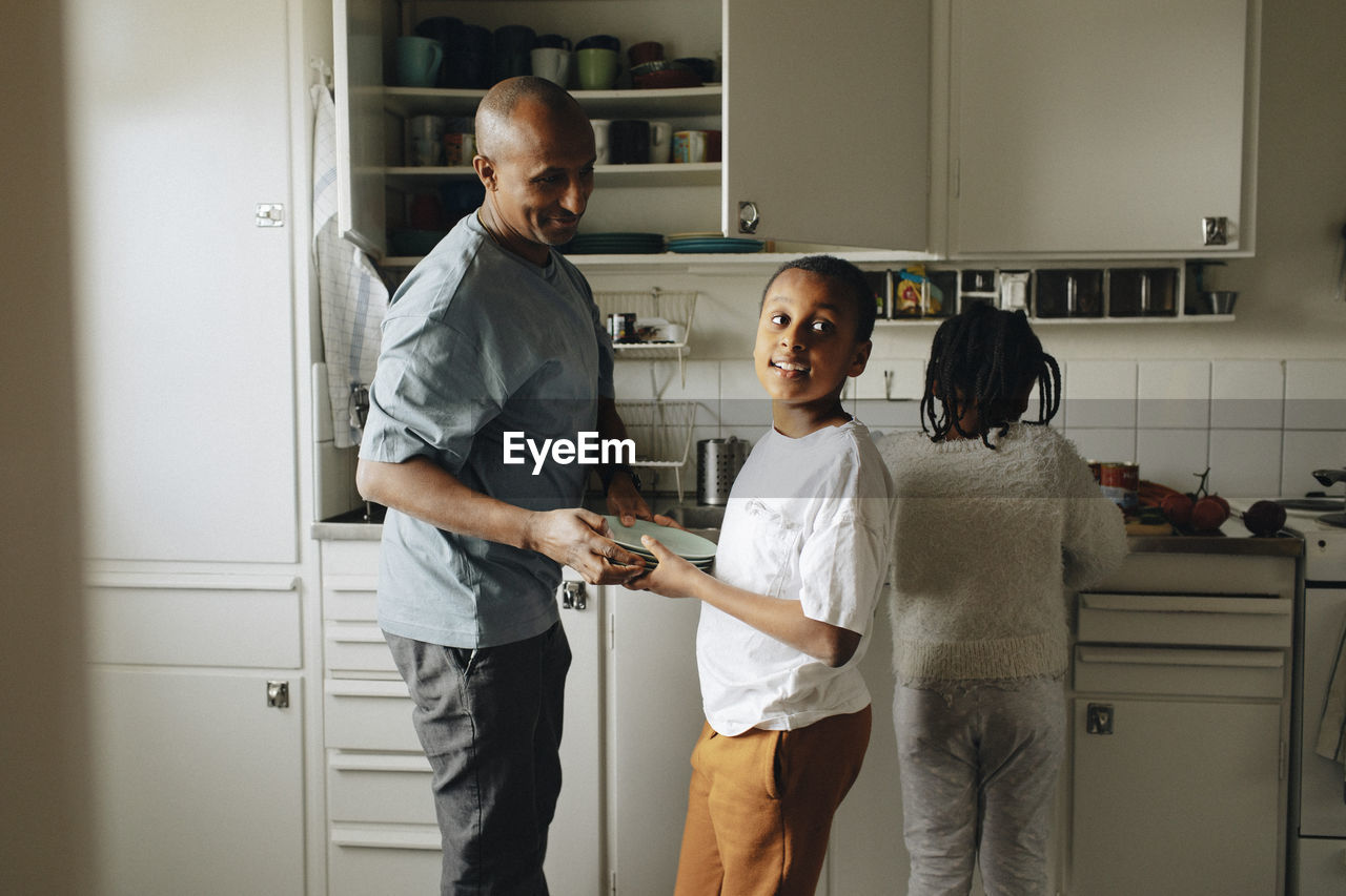 Son helping father while holding plates in kitchen at home