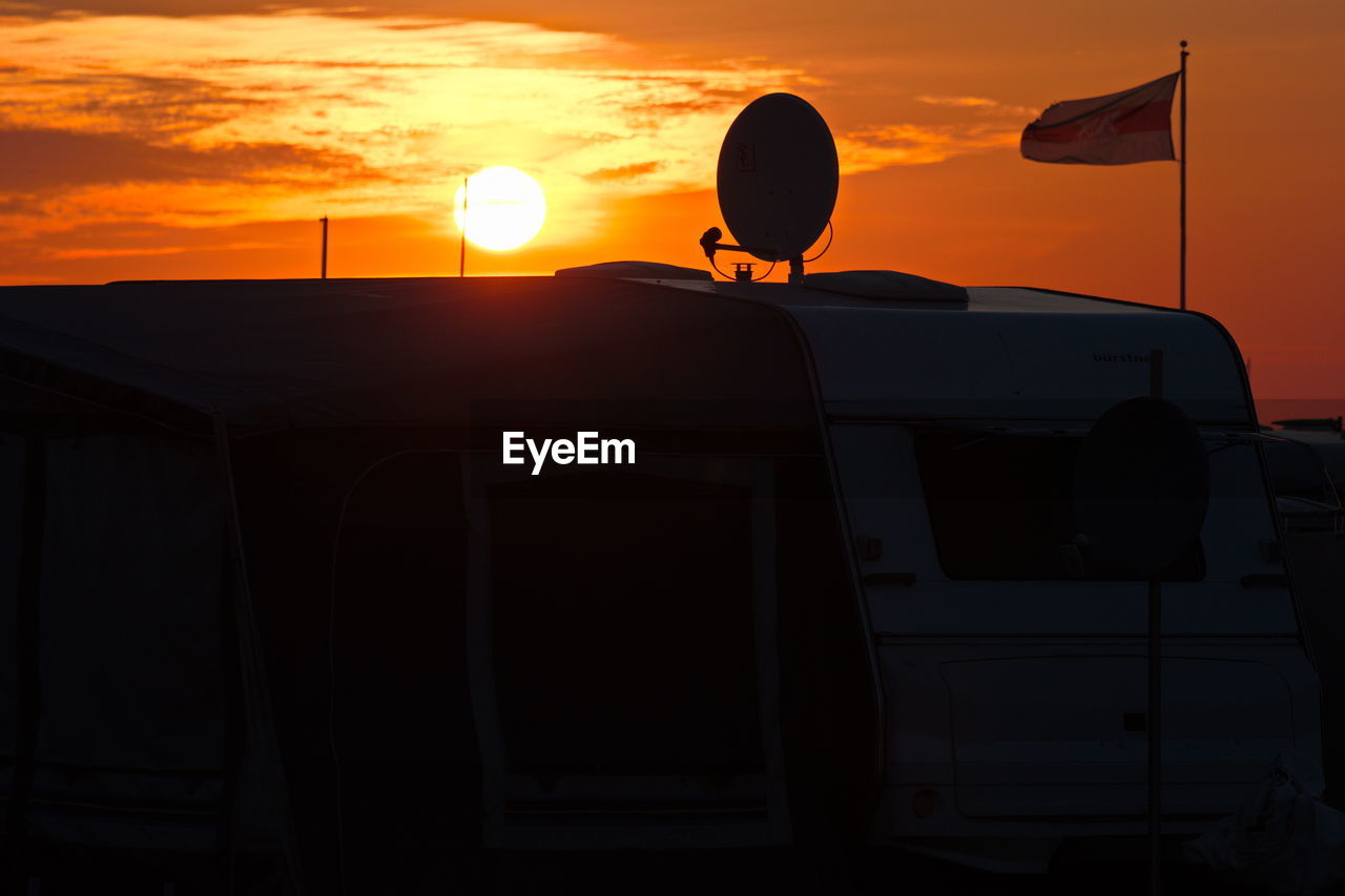 Flag and satellite dish on motor home against sky during sunset