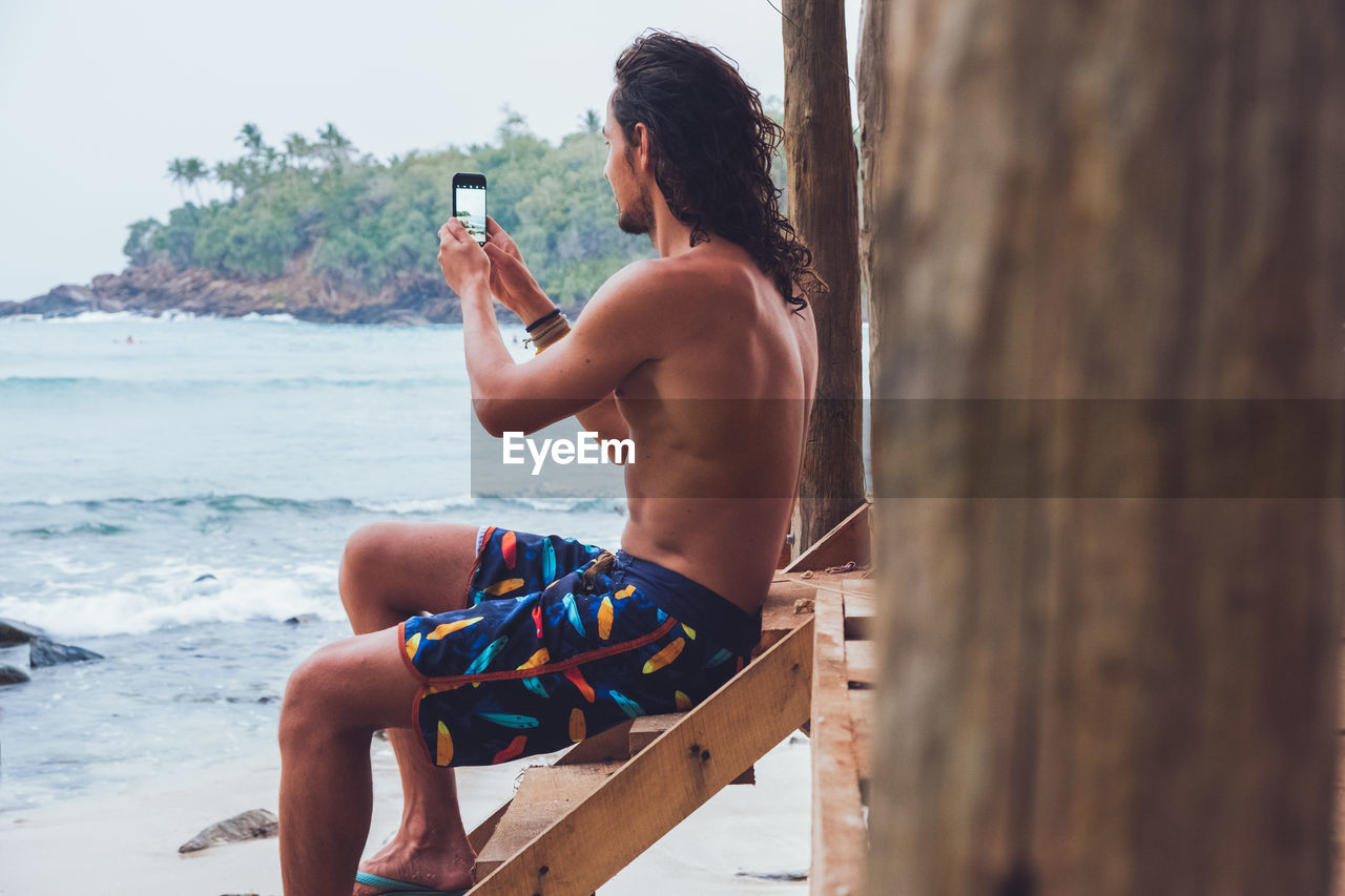 Side view of shirtless male tourist sitting on wooden terrace near sea and taking pictures with smartphone while spending holidays in hiriketiya in sri lanka