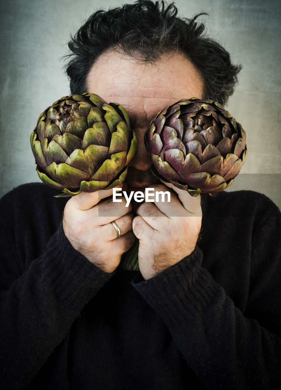 Close-up of man covering eyes with artichokes by wall