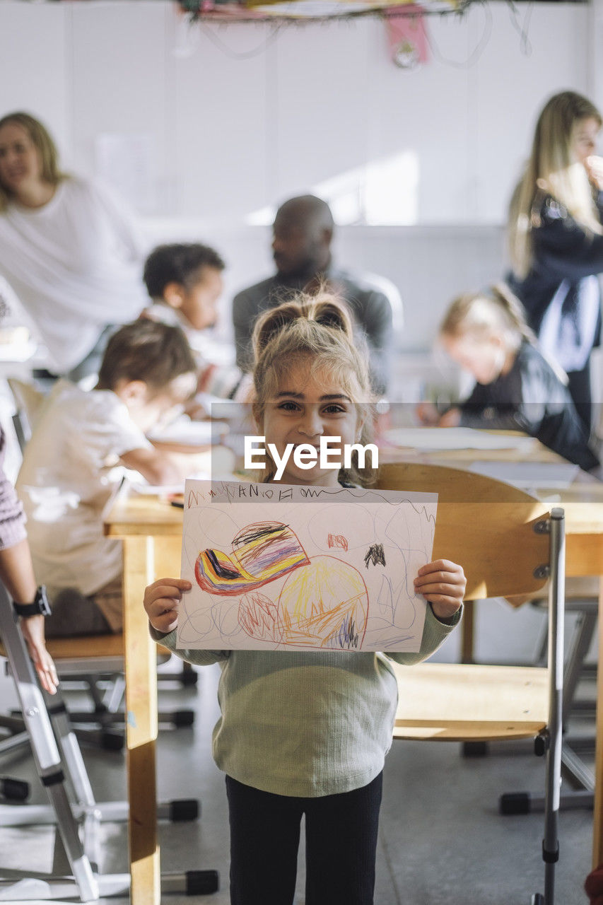 Portrait of smiling girl showing drawing while standing near bench in classroom at kindergarten