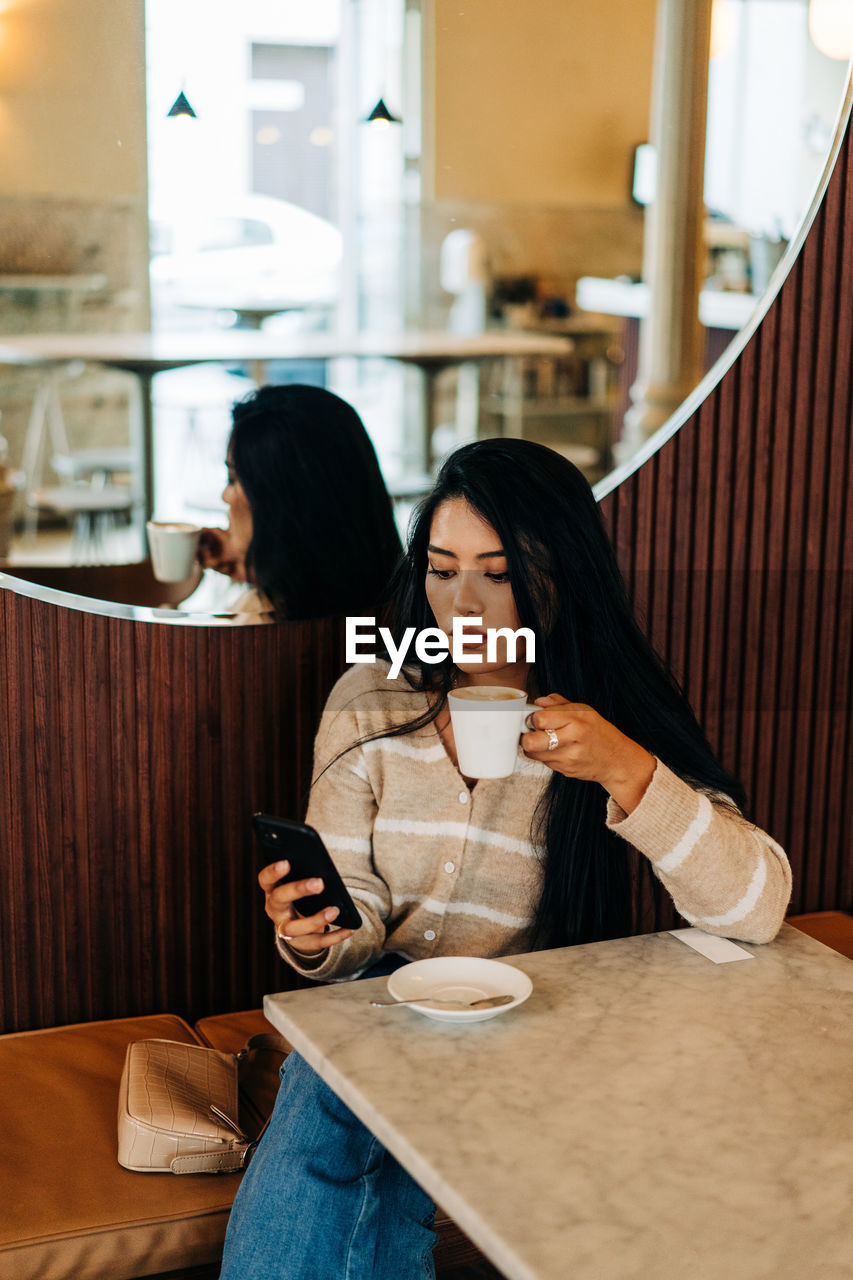 Young ethnic female with cup of hot drink text messaging on cellphone at cafeteria table against mirror