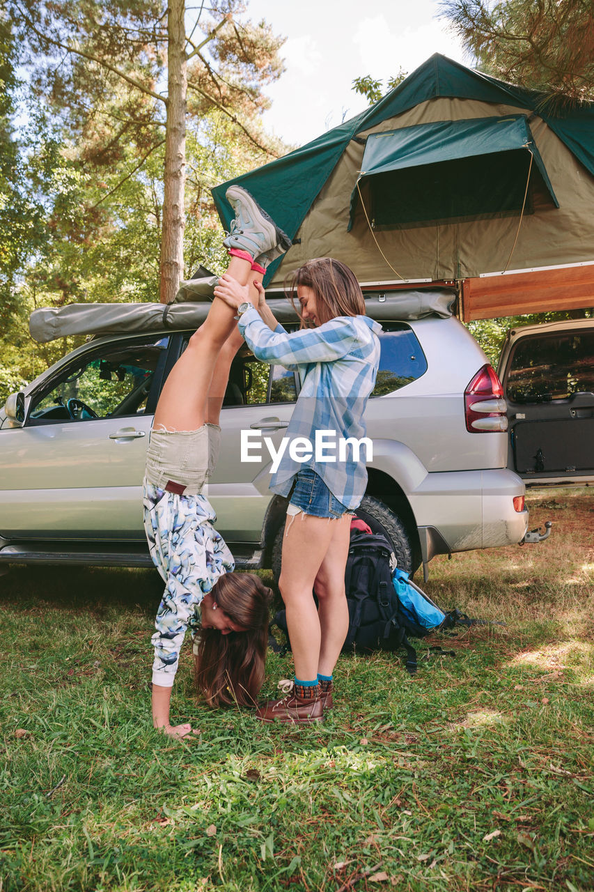 Woman helping friend in doing handstand by car at forest