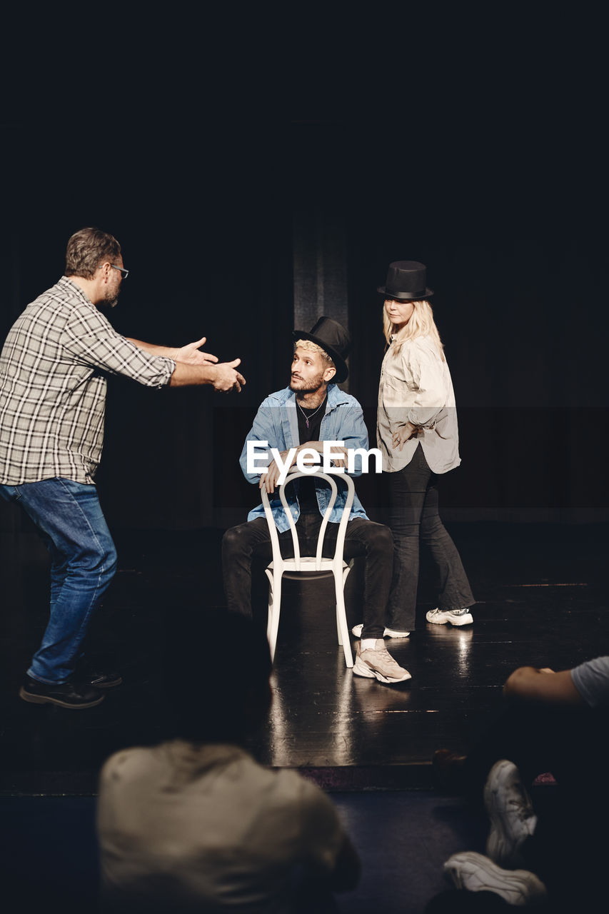 Full length of man guiding artist sitting on chair while rehearsing with woman on stage