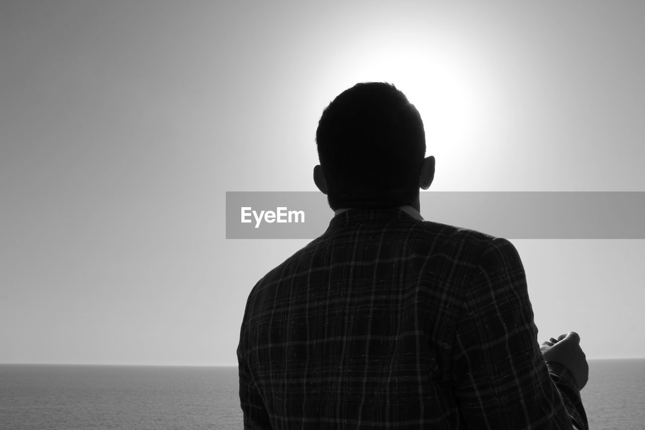 REAR VIEW OF MAN STANDING AT SEA AGAINST CLEAR SKY