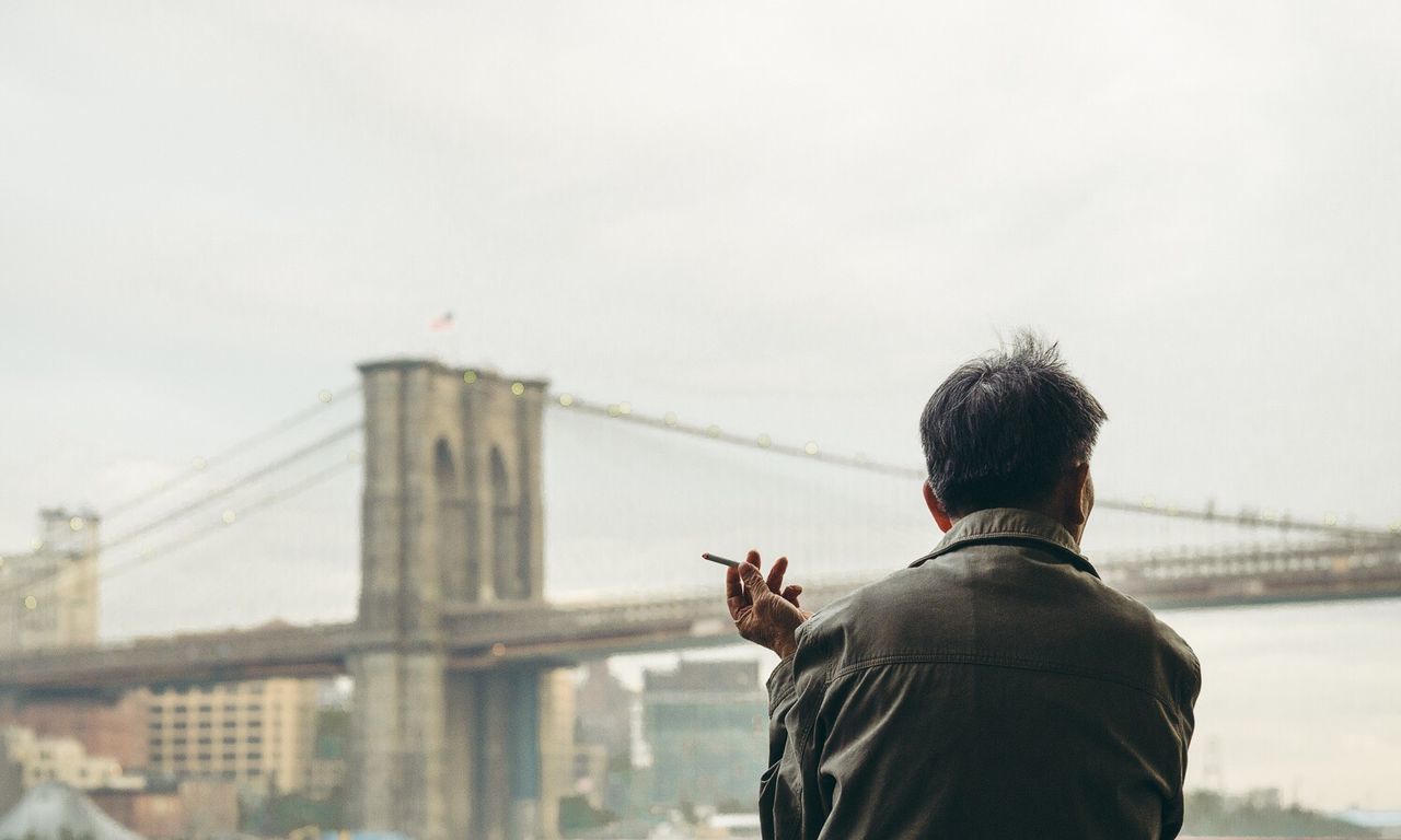 Rear view of man holding cigarette against brooklyn bridge