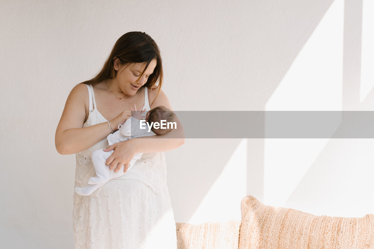 Adult woman embracing and pressing sleeping infant to chest gently against white wall