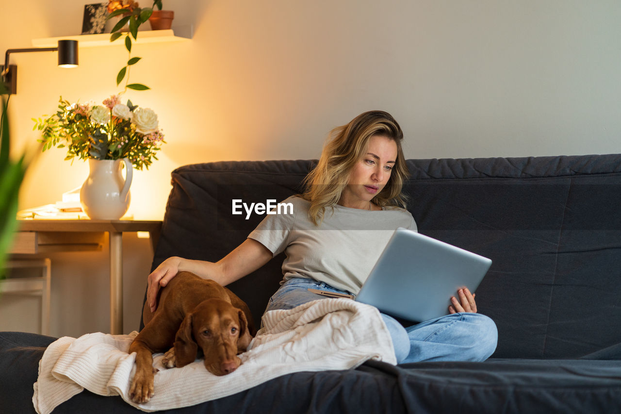 Woman sitting at couch, stroking her dog and working on laptop, surfing web at home in living room