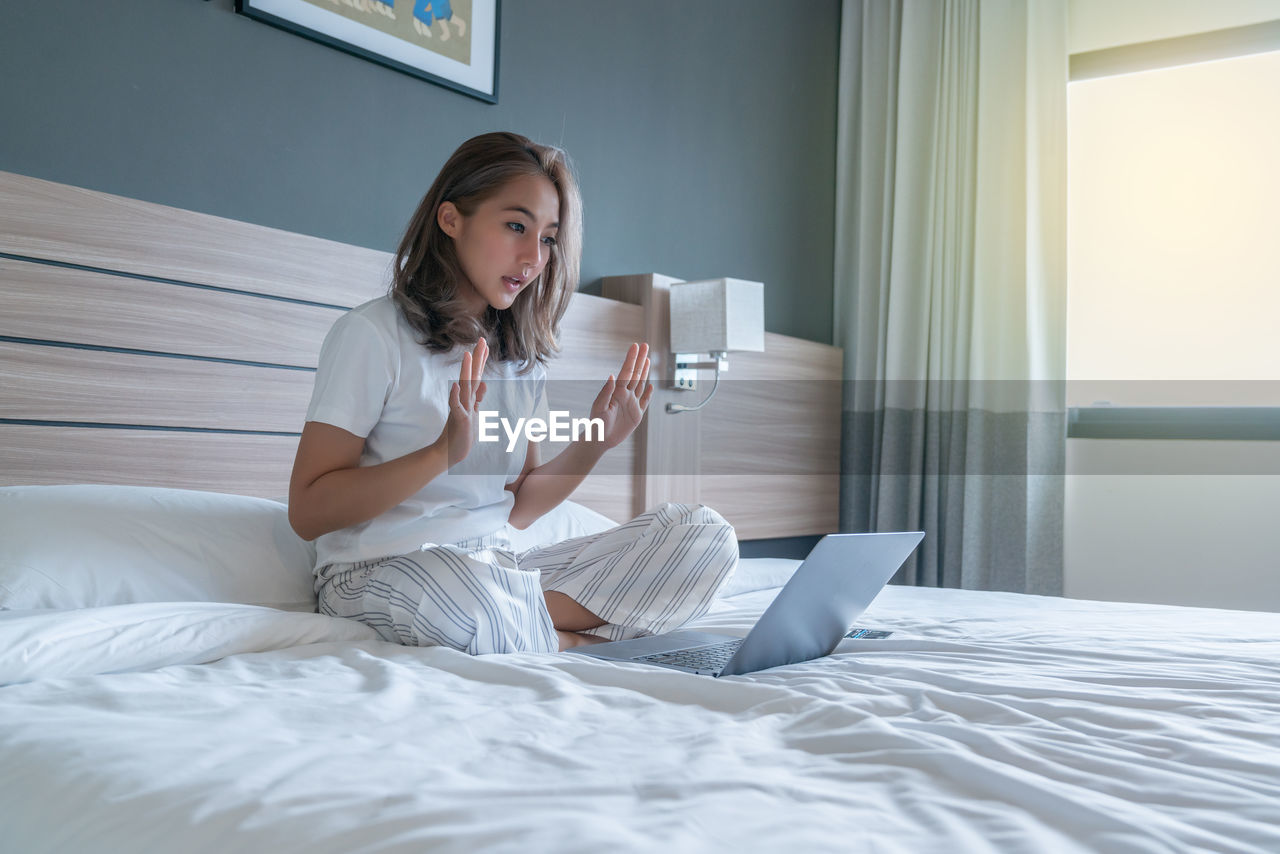YOUNG WOMAN USING PHONE WHILE SITTING ON BED AT HOME