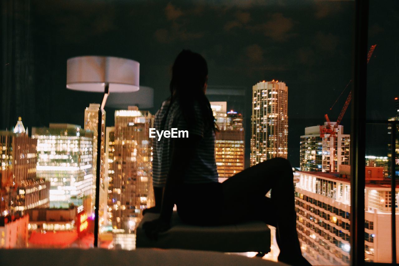 Woman on seat looking at illuminated buildings seen through window during night
