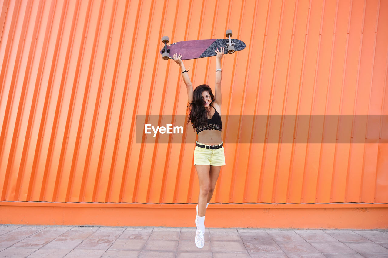 Portrait of young woman standing in front of corrugated iron wall with longboard in hands