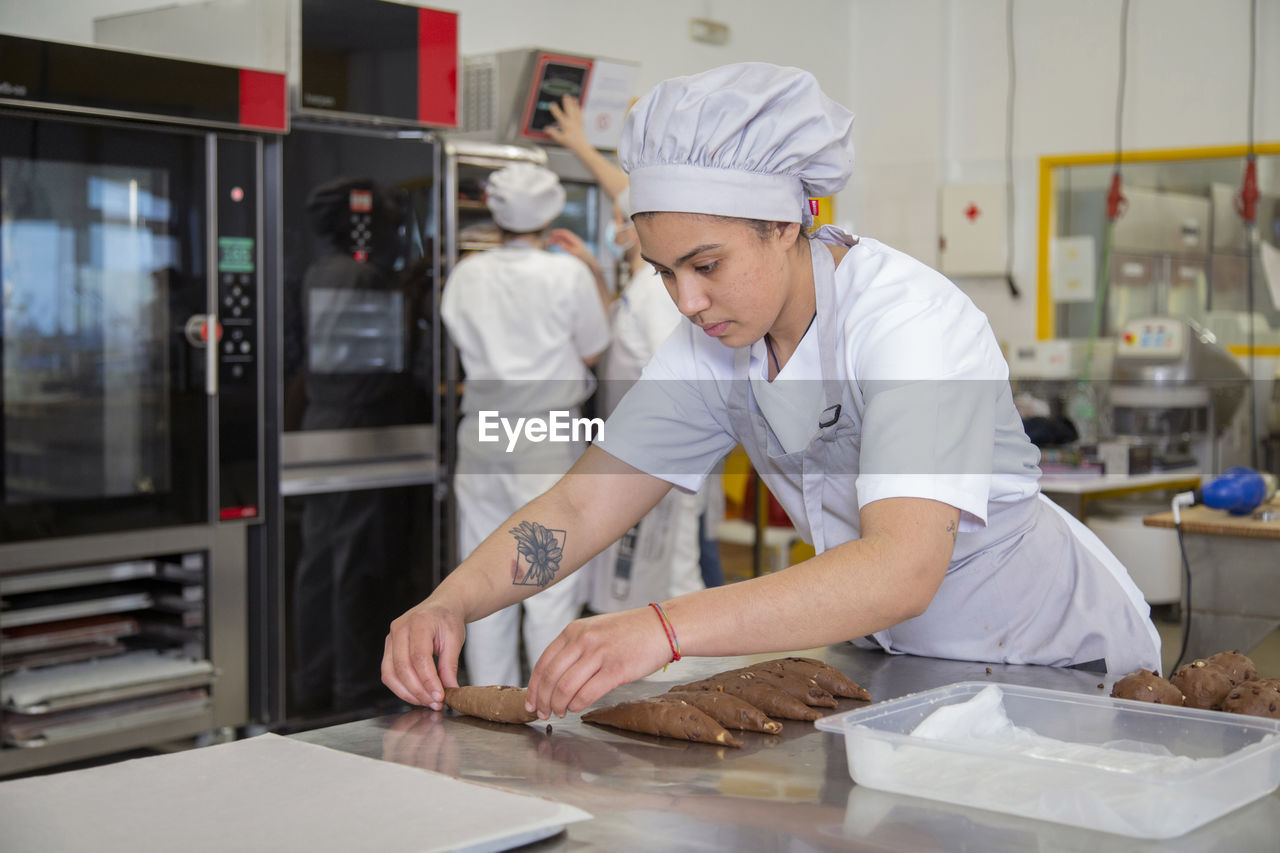 Focused female cook in uniform forming sweet dough for cookies on metal table while learning baking in light bakery school