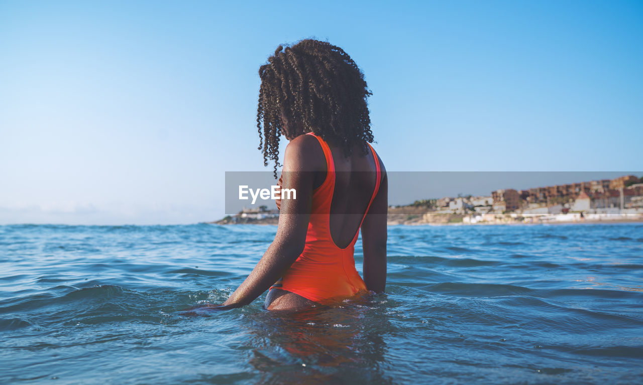 Side view of unrecognizable african american female tourist with curly hair in stylish red swimsuit standing in waving sea against cloudless blue sky