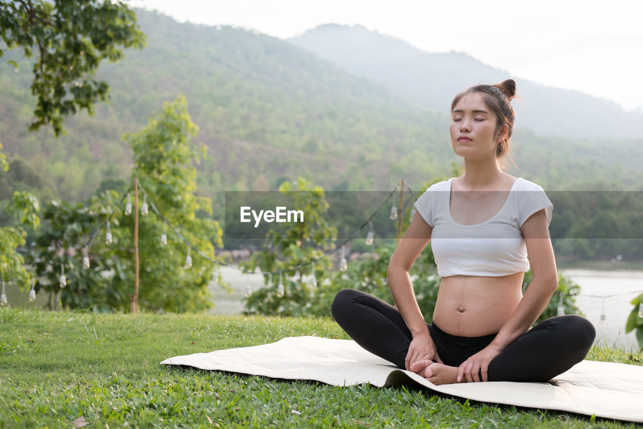 Pregnant woman with arms raised exercising while sitting on mat over grass against sky