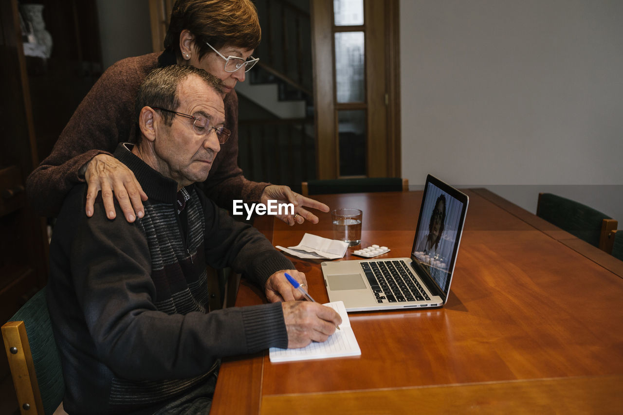 Senior man with social worker writing while video consultation with female doctor at home