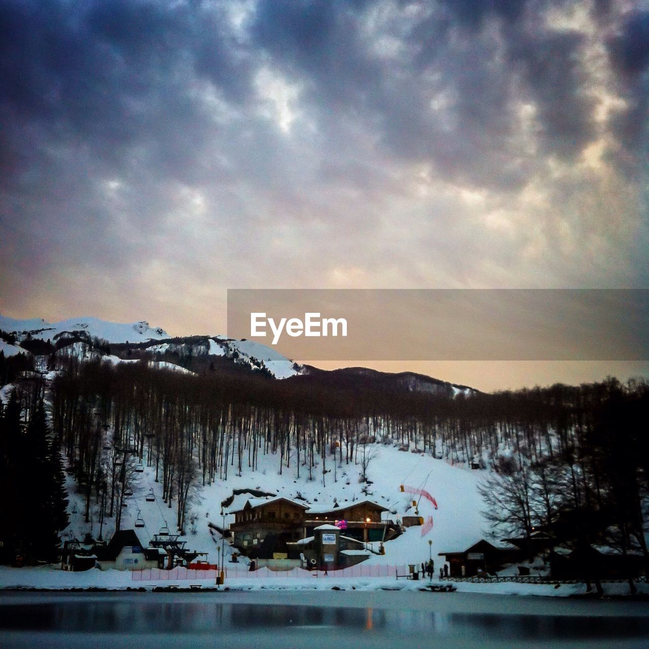 Frozen lake in front of ski resort on mountain against cloudy sky