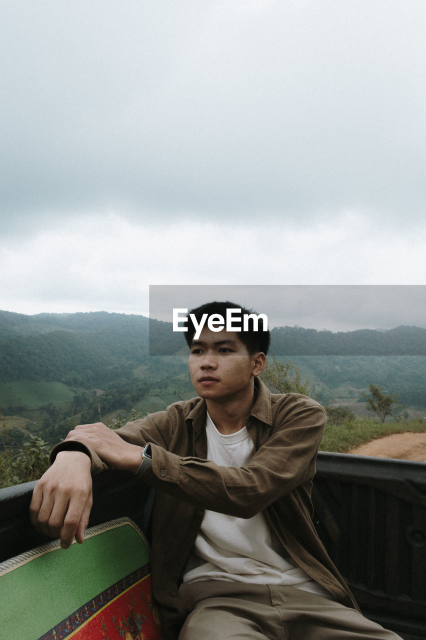 Young man sitting on car trunk against sky