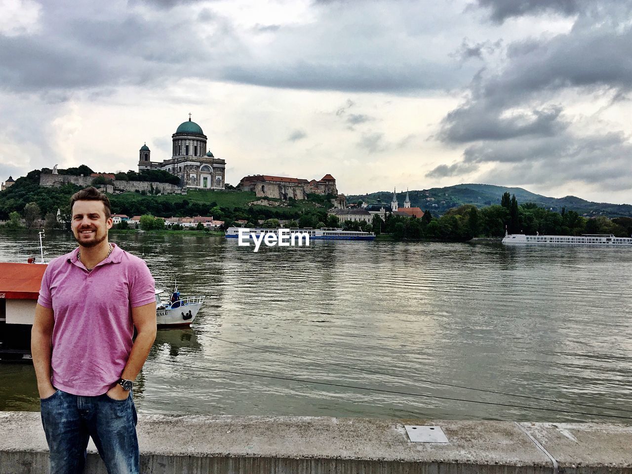 Man with hands in pockets standing by lake against cloudy sky at esztergom basilica