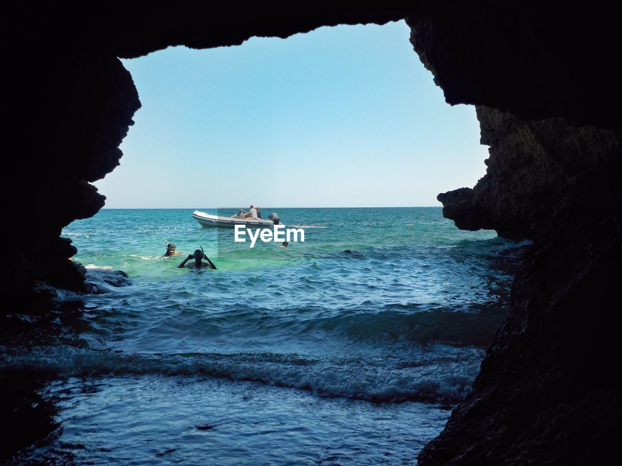 People swimming in sea seen through cave against clear sky