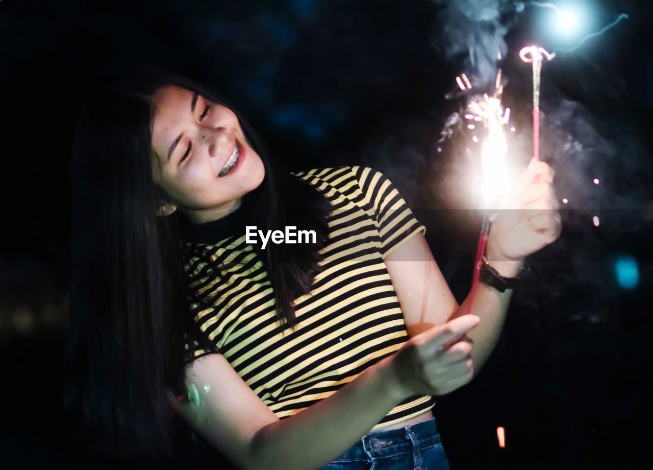 Smiling young woman burning sparklers while standing outdoors at night