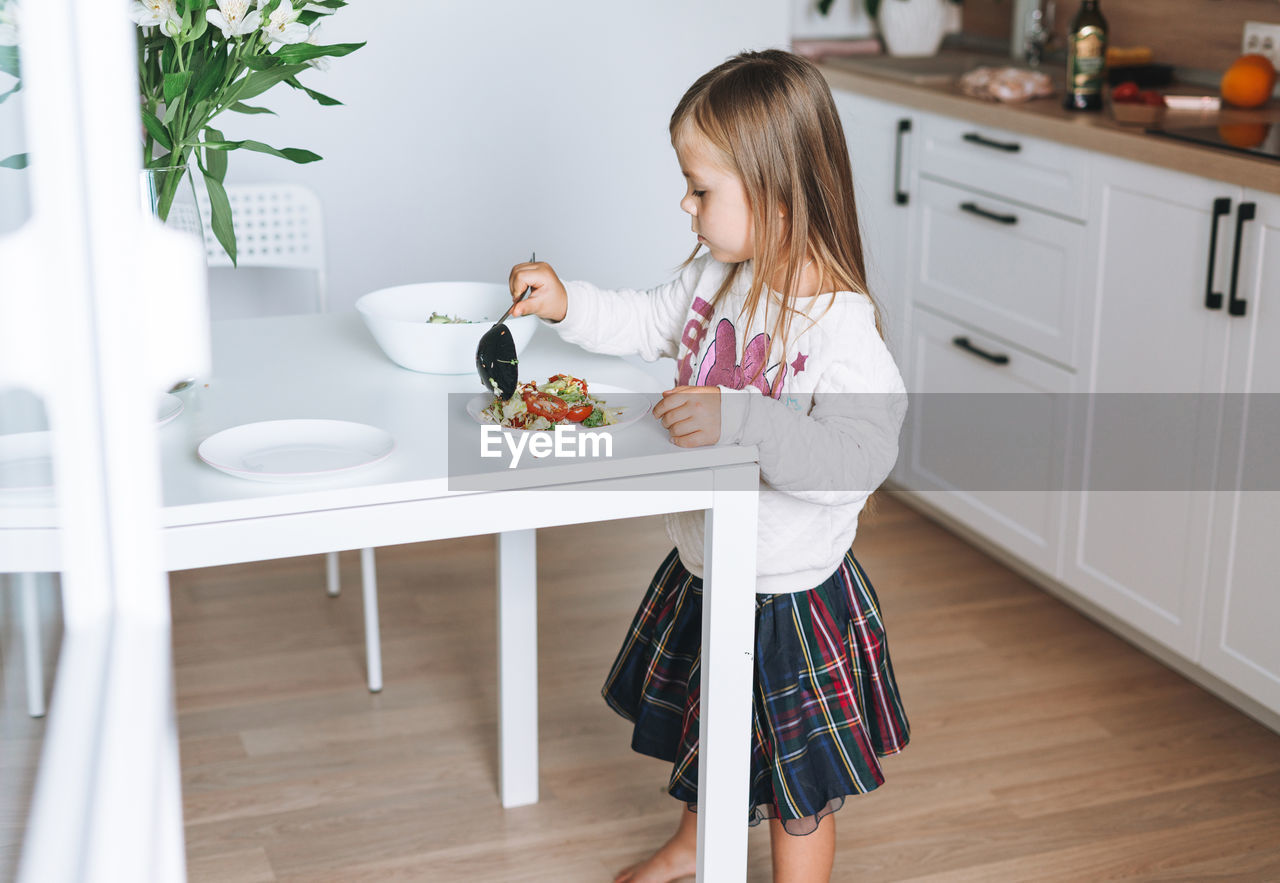 Pretty little girl with long hair puts on vegetable salad in kitchen with bright interior at home