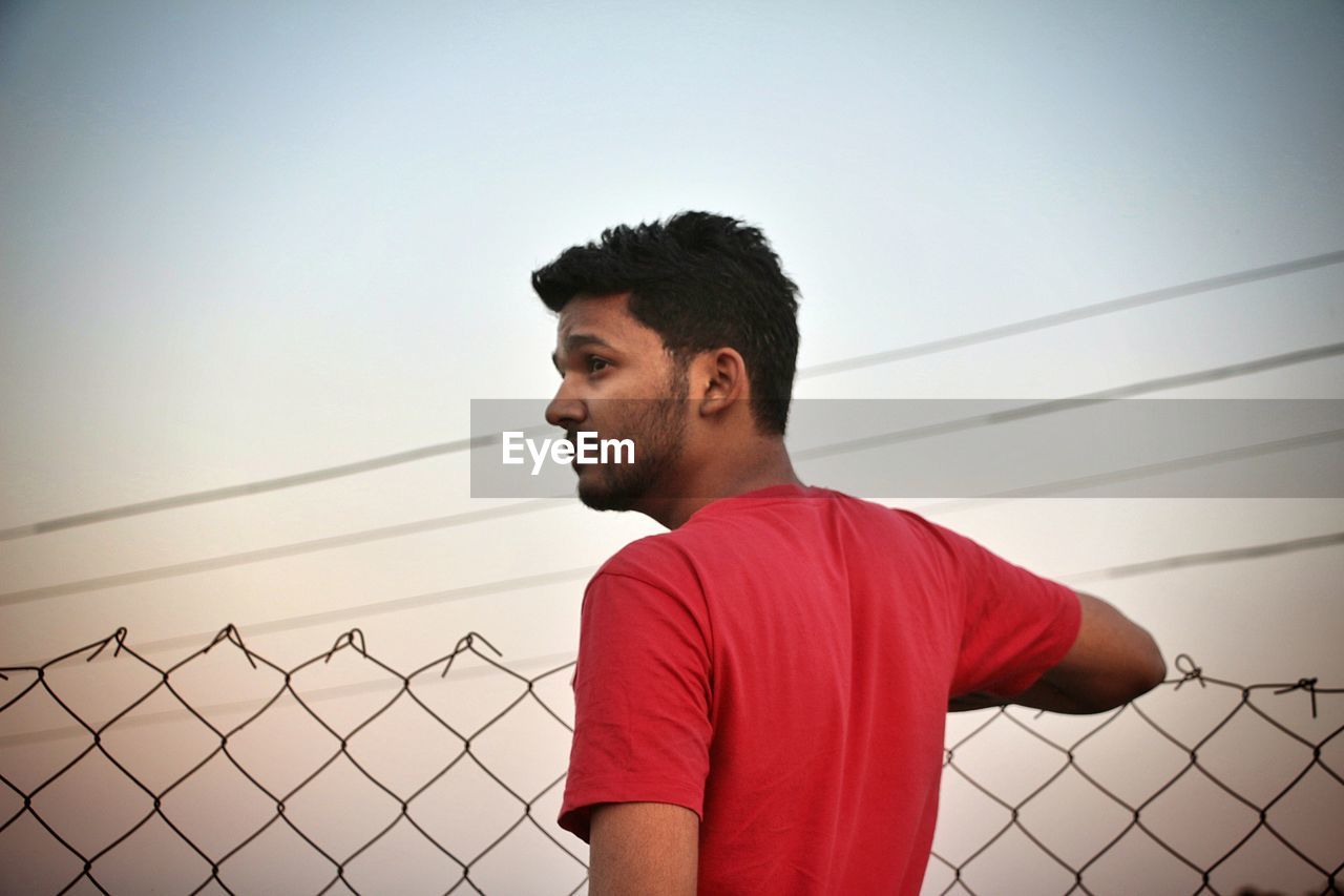 Thoughtful young man standing by chainlink fence against clear sky