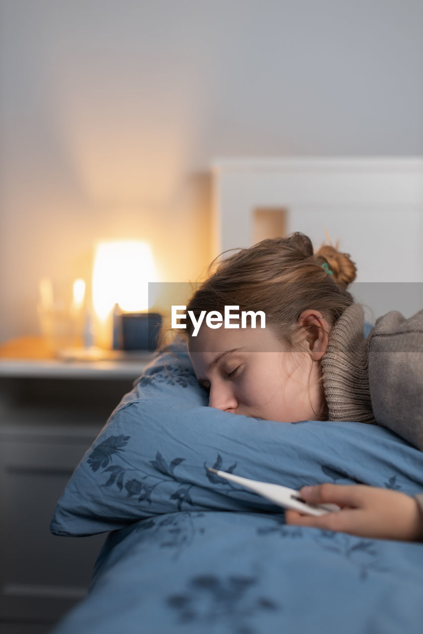 Young woman holding thermometer resting on bed at home