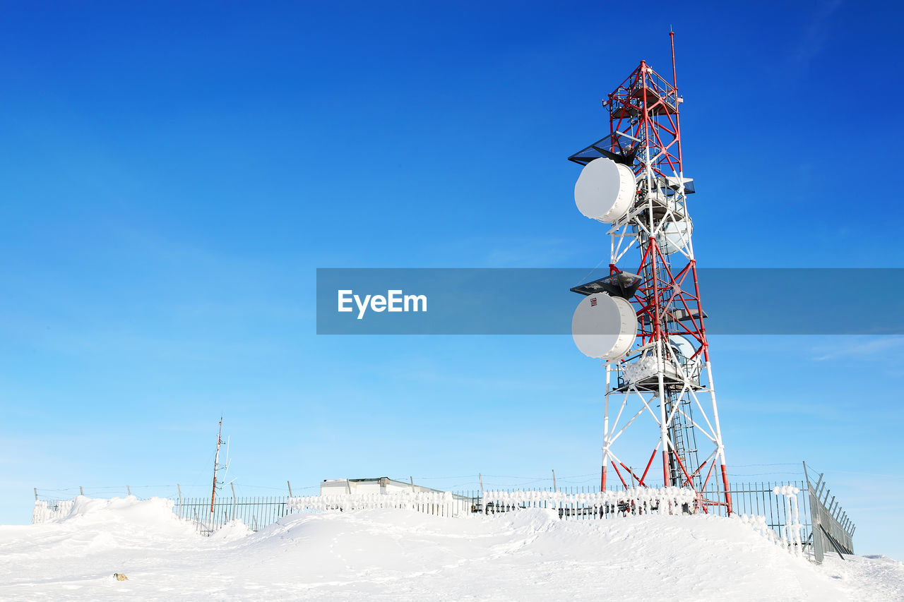 Low angle view of communication tower on snow field against blue sky
