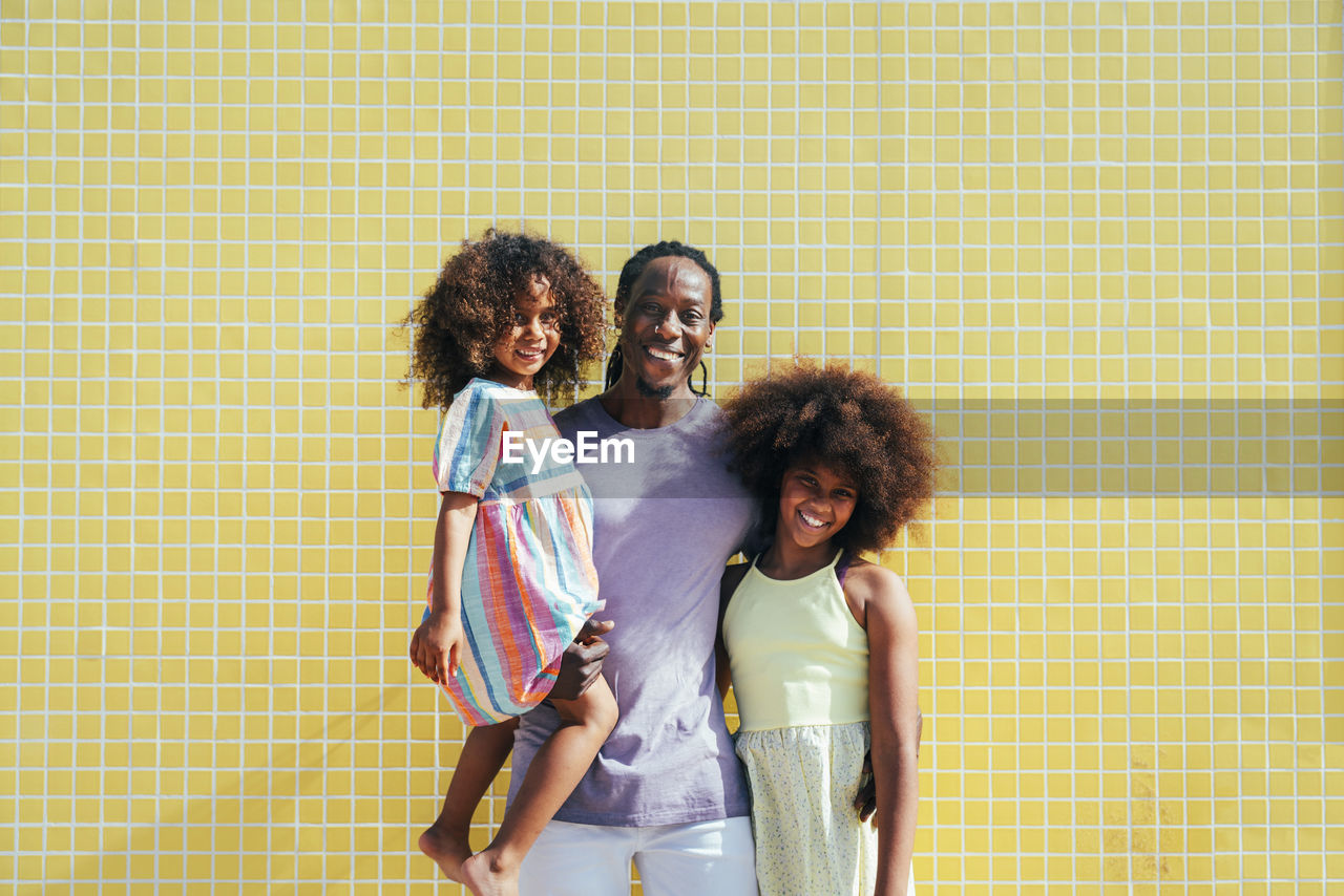Smiling father with daughters standing in front of yellow wall