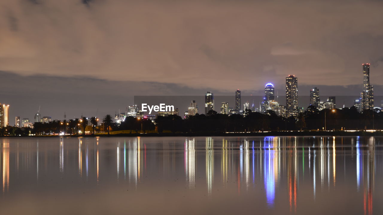 Albert park lake by illuminated buildings against cloudy sky at night