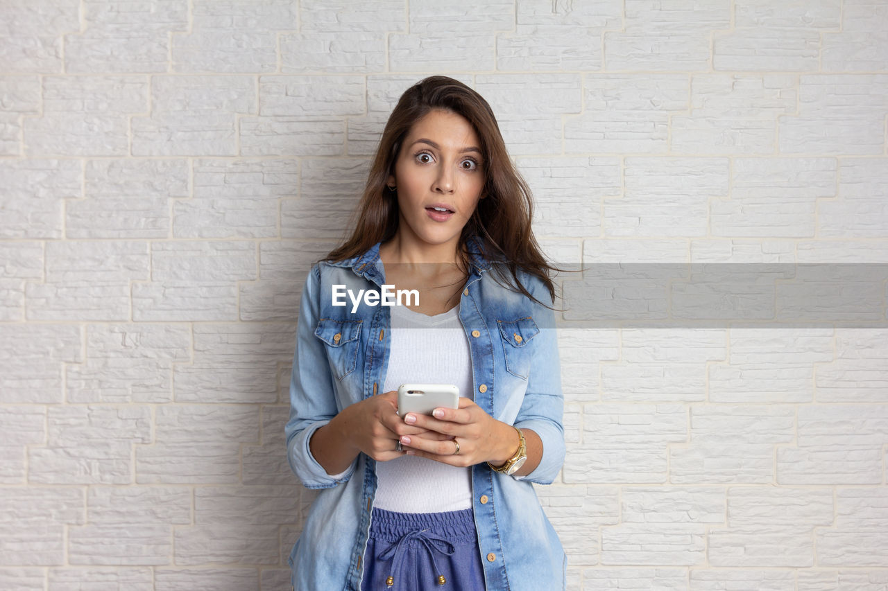 PORTRAIT OF BEAUTIFUL YOUNG WOMAN USING PHONE AGAINST WALL