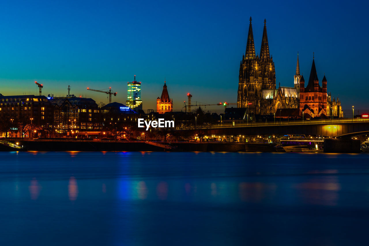 Illuminated buildings at waterfront against blue sky