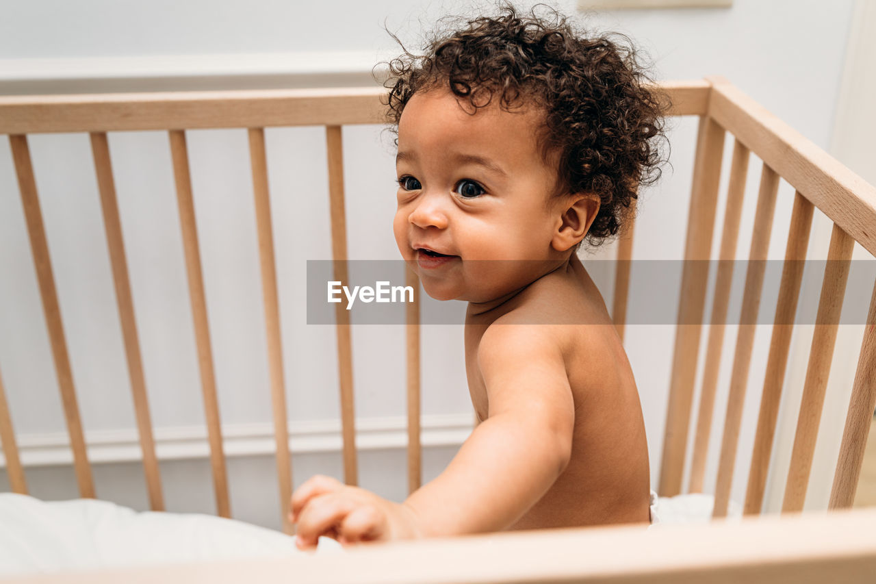 Cute baby boy looking away while sitting in crib at home