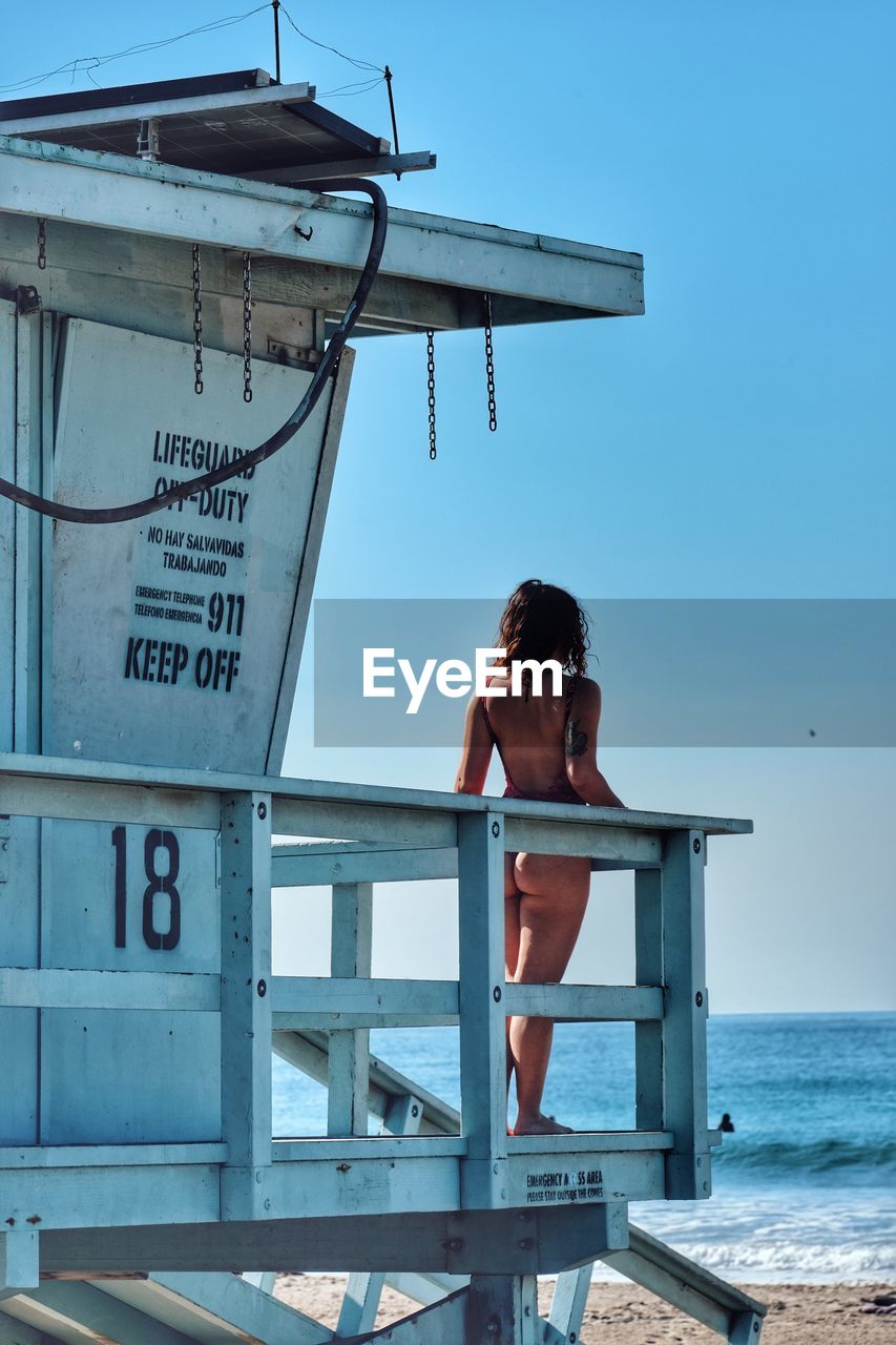 Rear view of seductive woman in bikini standing on lifeguard hut