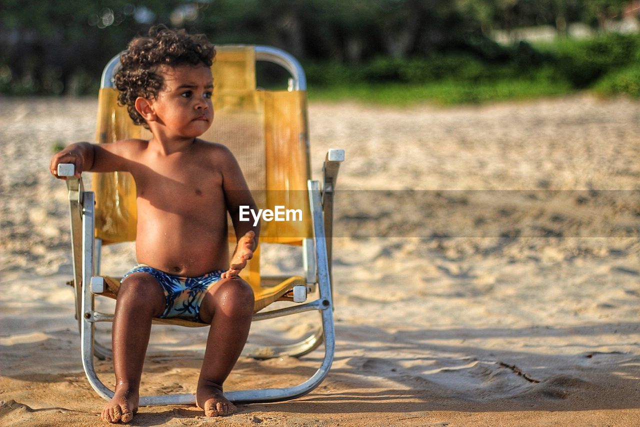 Full length of shirtless baby boy sitting on chair at beach